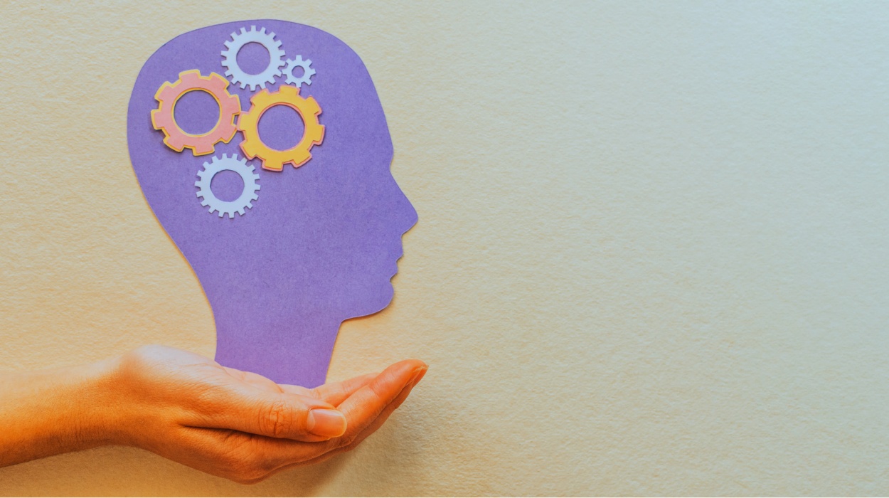 A hand seeming to hold up a paper cut-out illustration of a human head with gears inside