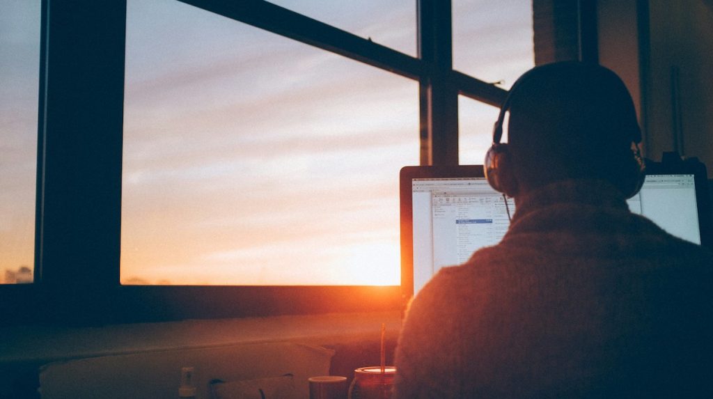 Man with headphones on, sitting in front of a laptop, with the sun setting through a window
