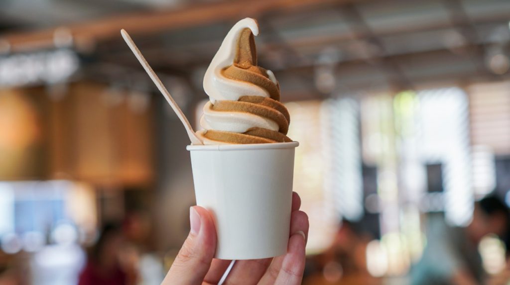 Soft-serve ice cream in a cup being held up in front of a busy office-like background