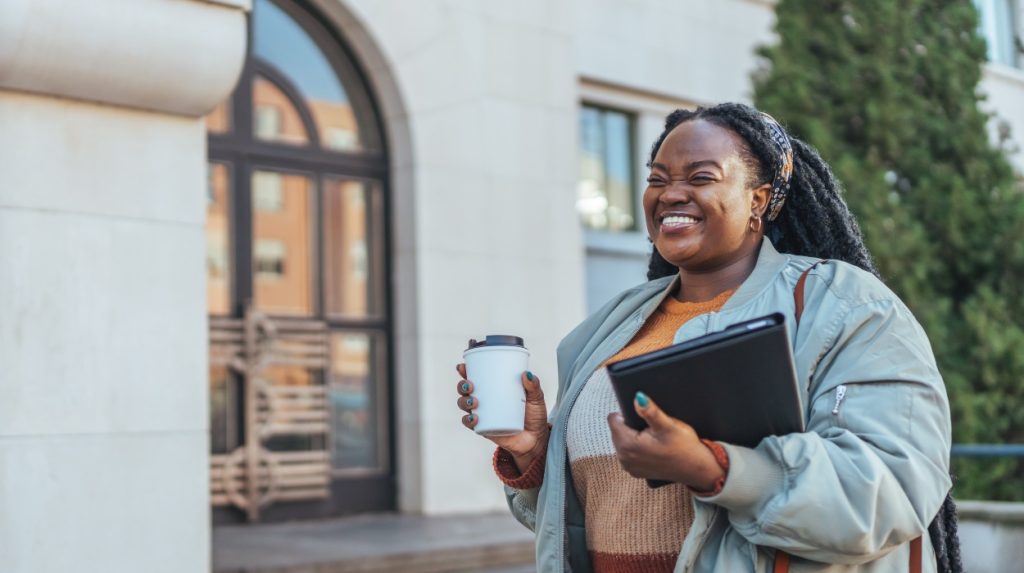Smiling woman holding a to-go coffee cup and a laptop outside of an office building