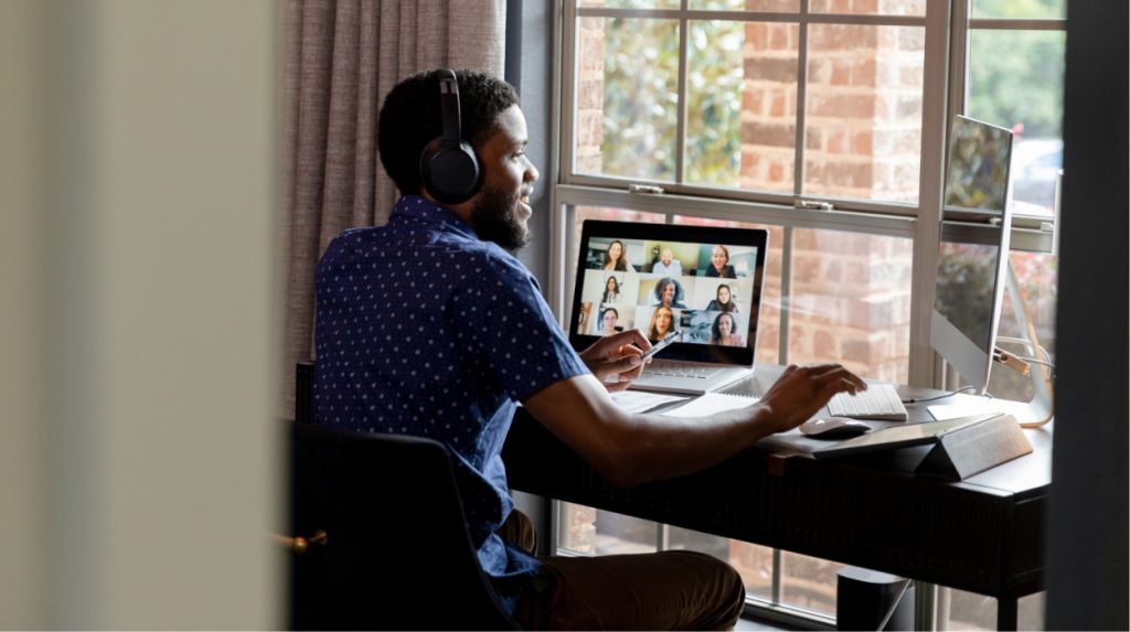 Man sitting at a desk with headphones on and a laptop in front of him with a virtual meeting displayed