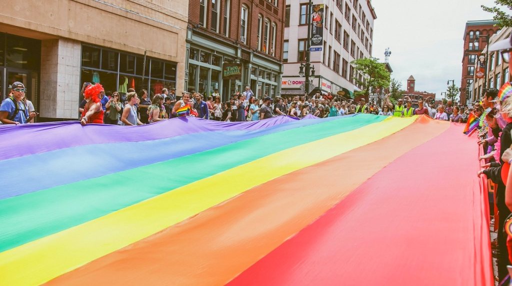 Giant rainbow Pride flag being held up by dozens of people in a city street